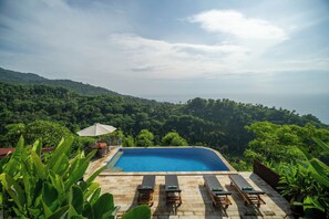 A swimming pool with lounge chairs and an umbrella, surrounded by lush greenery and hills.