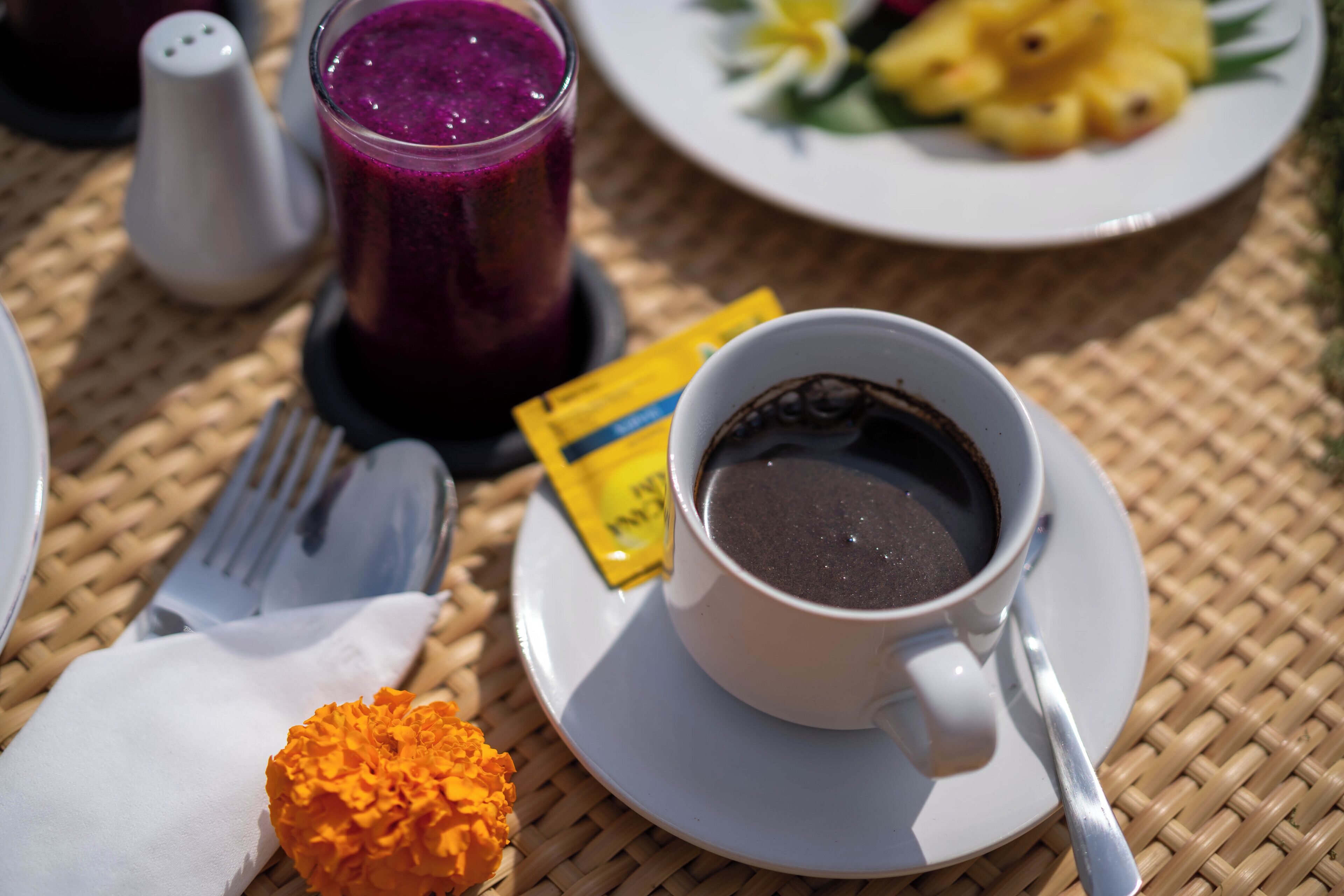 A table with a cup of coffee, a glass of purple smoothie, a plate of fruit, a salt shaker, and a packet of sugar.