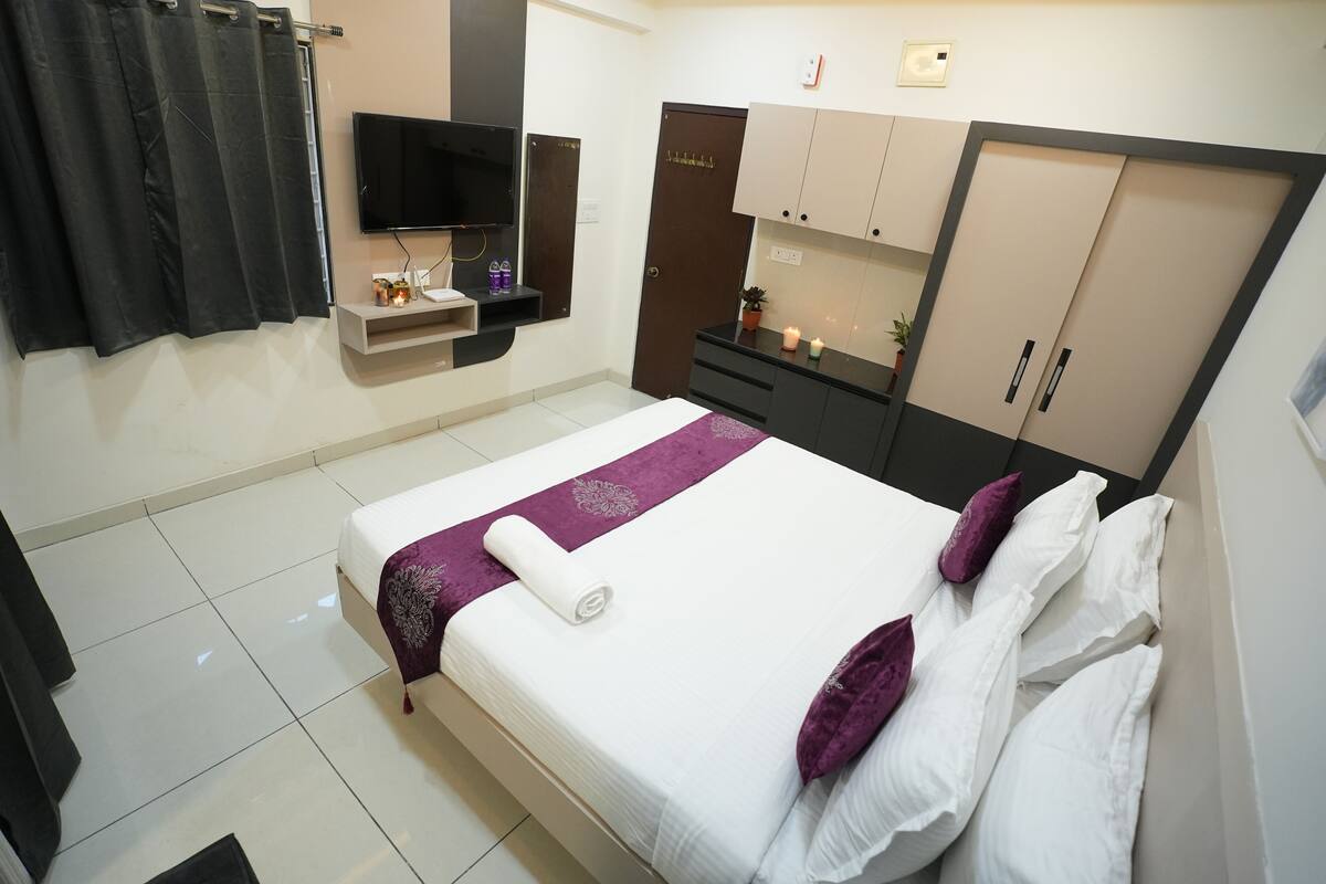 Une chambre d’hôtel bien rangée, avec un lit, une télévision, une armoire et une petite étagère contenant des articles de toilette.