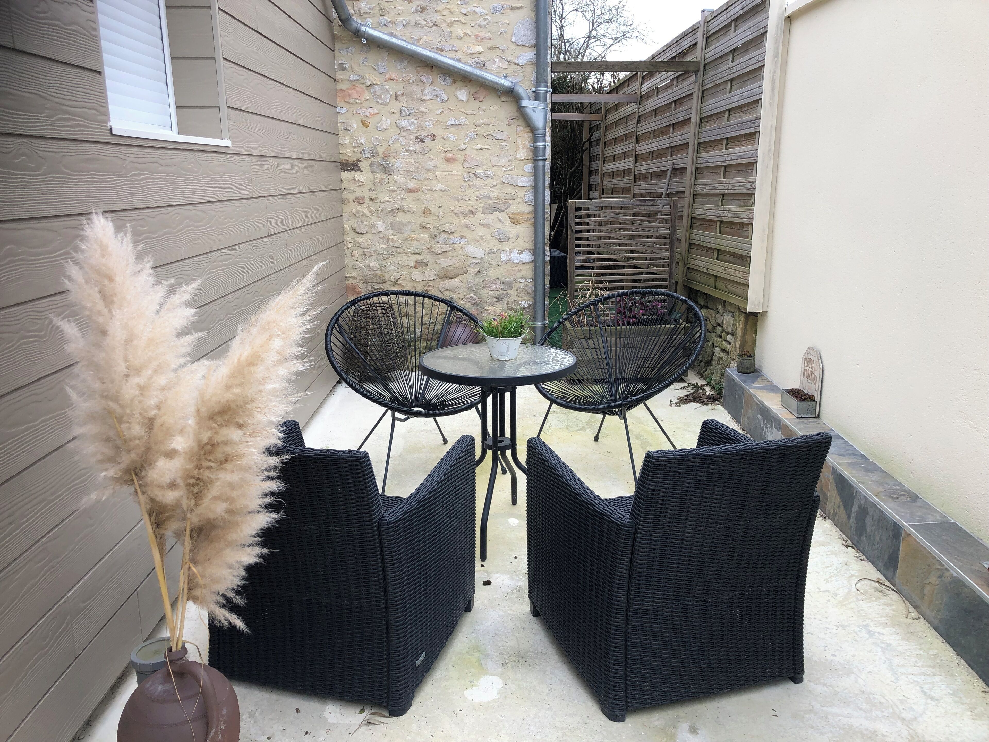 Une terrasse avec deux chaises en osier noir et une petite table ronde, un pot de fleurs et des herbes des pampas.