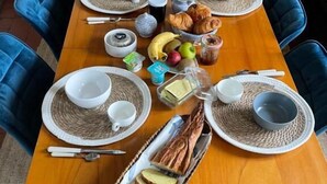 Egy fából készült étkezőasztal, melyen tányérok, tálak, evőeszközök, kenyér, gyümölcsök és különféle asztali fűszerek találhatók.