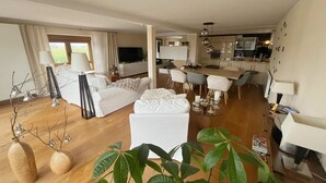 Una sala moderna con sofá, reposapiés y comedor con sillas. Se observa una cocina al fondo, con televisión y armarios.