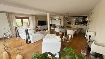 Ein modernes Wohnzimmer mit Sofa, Hocker und Essbereich mit Stühlen. Im Hintergrund ist eine Küche mit Fernseher und Schränken zu sehen.