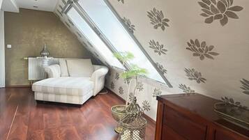 Ein Zimmer mit schrägverdeckter Decke, einem weißen Sofa, einem Holzschrank und einer Zimmerpflanze im Metalltopf.