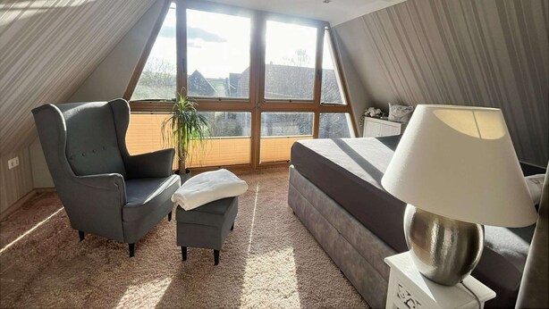 Un dormitorio moderno con un ventanal grande, un sillón gris, un pequeño reposapiés, un sofá y una lámpara de noche.