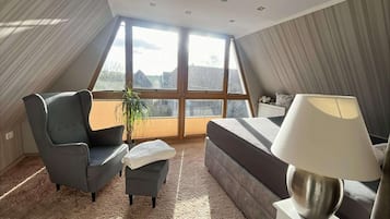 Ein modernes Schlafzimmer mit großem Fenster, einem grauen Sessel, einem kleinen Hocker, einem Sofa und einer Nachttischlampe.