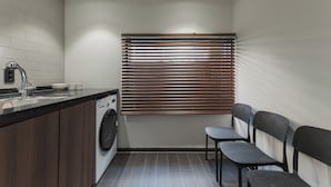 A compact kitchen with a washing machine, a sink, and a window with blinds. - J nine Lodge (Seoul)