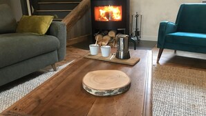 A cozy living room with a wood-burning stove, a grey sofa, a teal armchair, and a round wooden table with cups and a kettle.