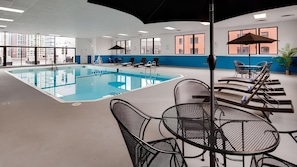 A swimming pool with lounge chairs and tables, large windows, and a view of buildings outside.