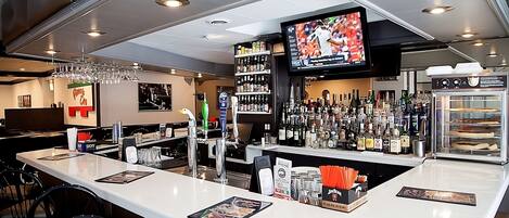 Zona de bar con mostrador, taburetes, una pantalla de televisión y una variedad de botellas y vasos.
