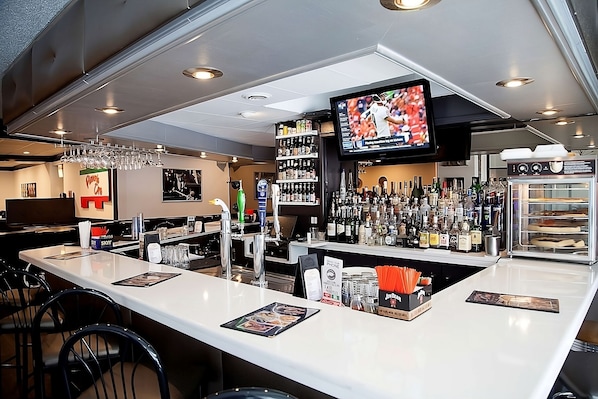 A bar area with a counter, bar stools, a television screen, and a variety of bottles and glasses.