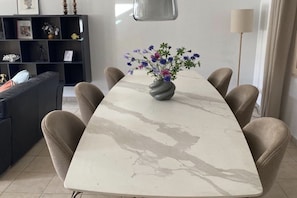 Un coin repas comprenant une table en marbre, six chaises, un vase avec des fleurs, un canapé, une bibliothèque et une lampe sur pied.