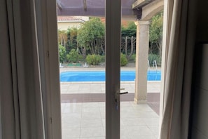 Une porte ouverte donne sur un espace piscine, d’où l’on peut admirer un jardin et un bâtiment en arrière-plan.