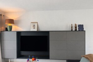 A modern living room with a large flat-screen TV mounted on a grey cabinet, a lamp, and decorative items on the mantel. - Detached Family Villa in Les Cammazes (Les Cammazes)
