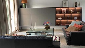 Un salon moderne avec une table basse en verre, un canapé en cuir noir, un fauteuil rembourré et une bibliothèque intégrée agrémentée d’objets décoratifs.