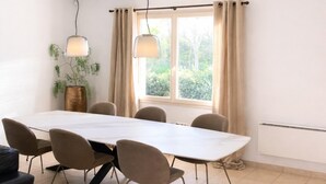 Un espace repas comprenant une longue table, six chaises, deux lampes suspendues, une plante en pot et une fenêtre avec des rideaux.