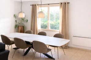 A dining area with a long table, six chairs, hanging pendant lights, a potted plant, and a window with curtains. - Detached Family Villa in Les Cammazes (Les Cammazes)