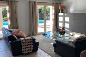 A living room with a sofa, armchair, glass coffee table, and a view of a swimming pool through large windows. - Detached Family Villa in Les Cammazes (Les Cammazes)