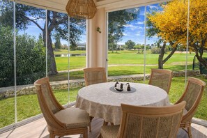 Une table ronde recouverte d’une nappe blanche, entourée de quatre chaises en osier, est placée sur un balcon d’où l’on peut admirer un terrain de golf et des arbres.