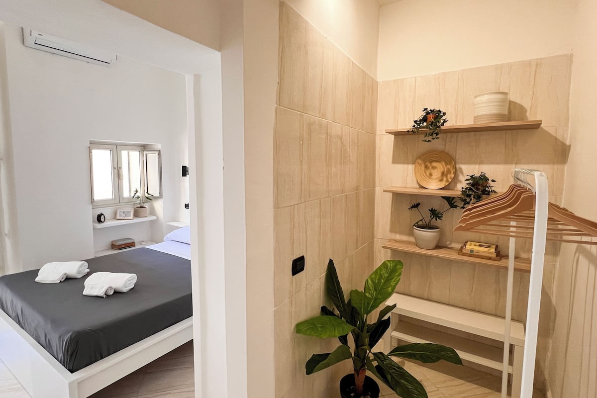 Une chambre à coucher comprenant un lit, une table de chevet, un miroir, une étagère avec des plantes et un porte-vêtements suspendu.