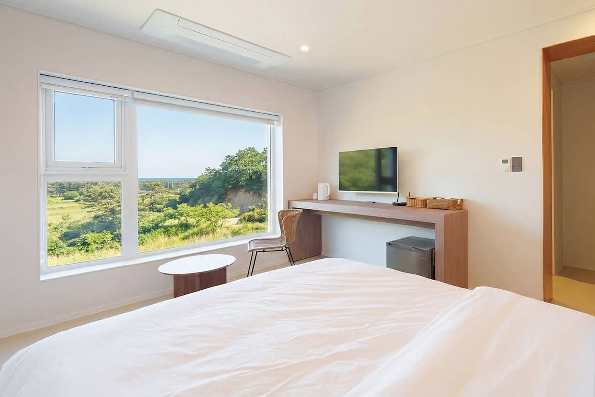 A modern hotel room with a large bed, a desk with a TV, and a view of a green landscape.