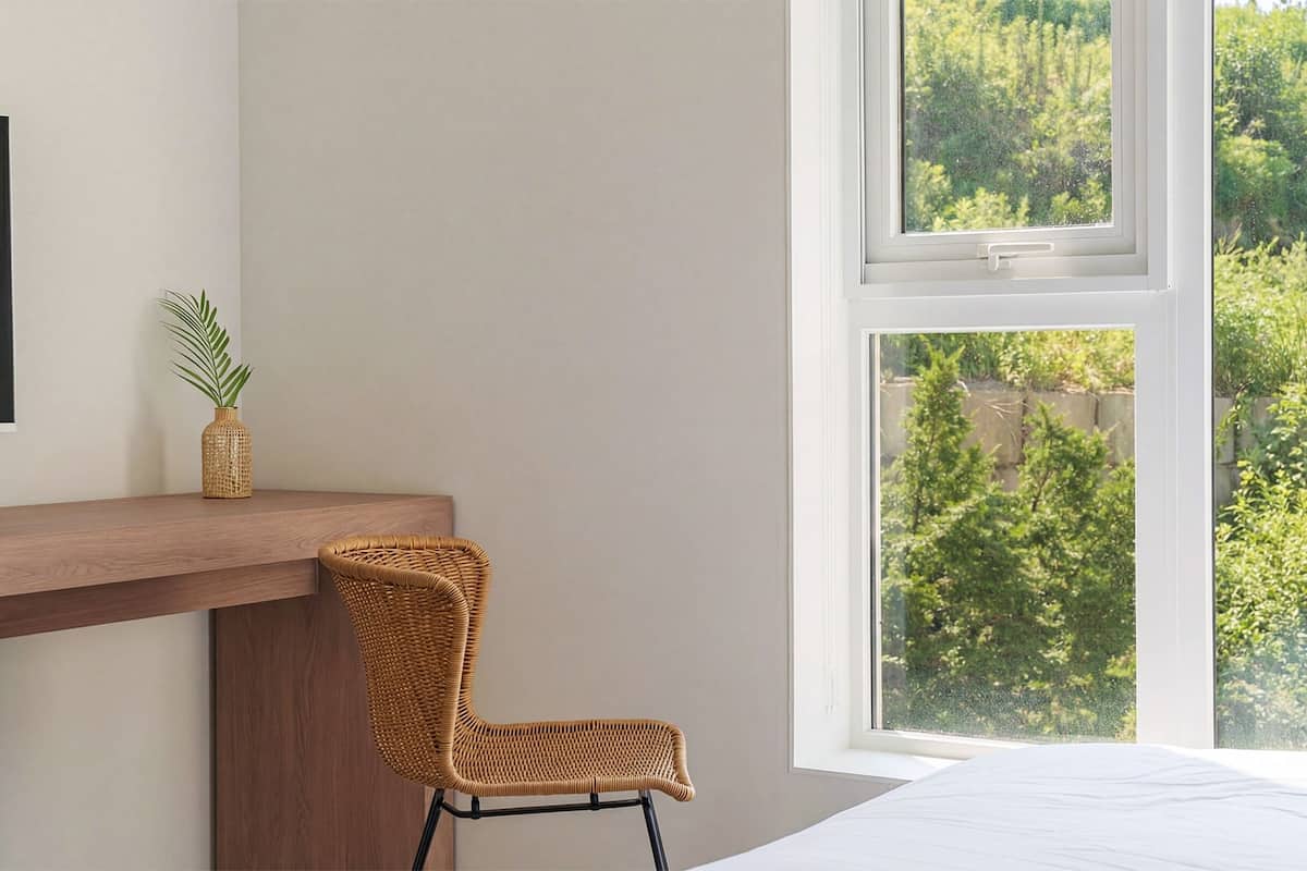 A bedroom with a large window, a wooden bedside table, a wicker chair, and a potted plant.