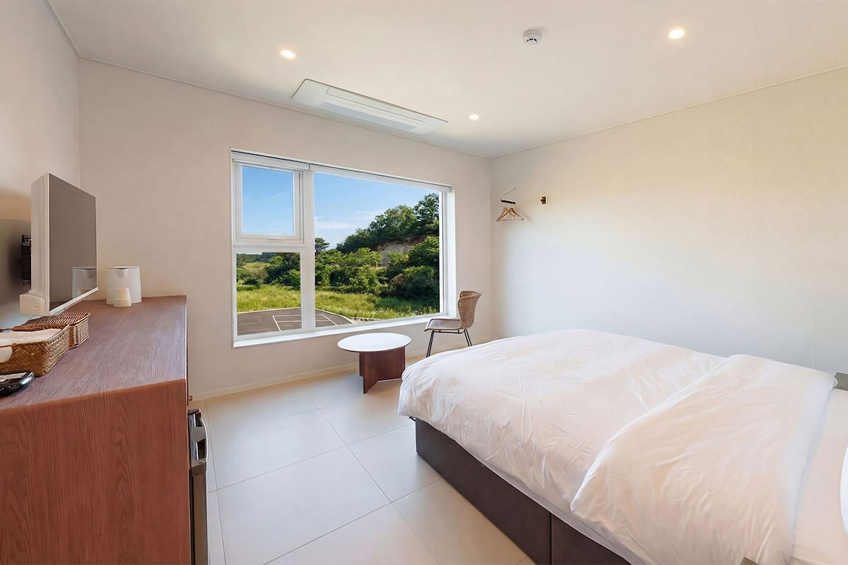 A bedroom with a large window, a bed, a bedside table, and a mirror.