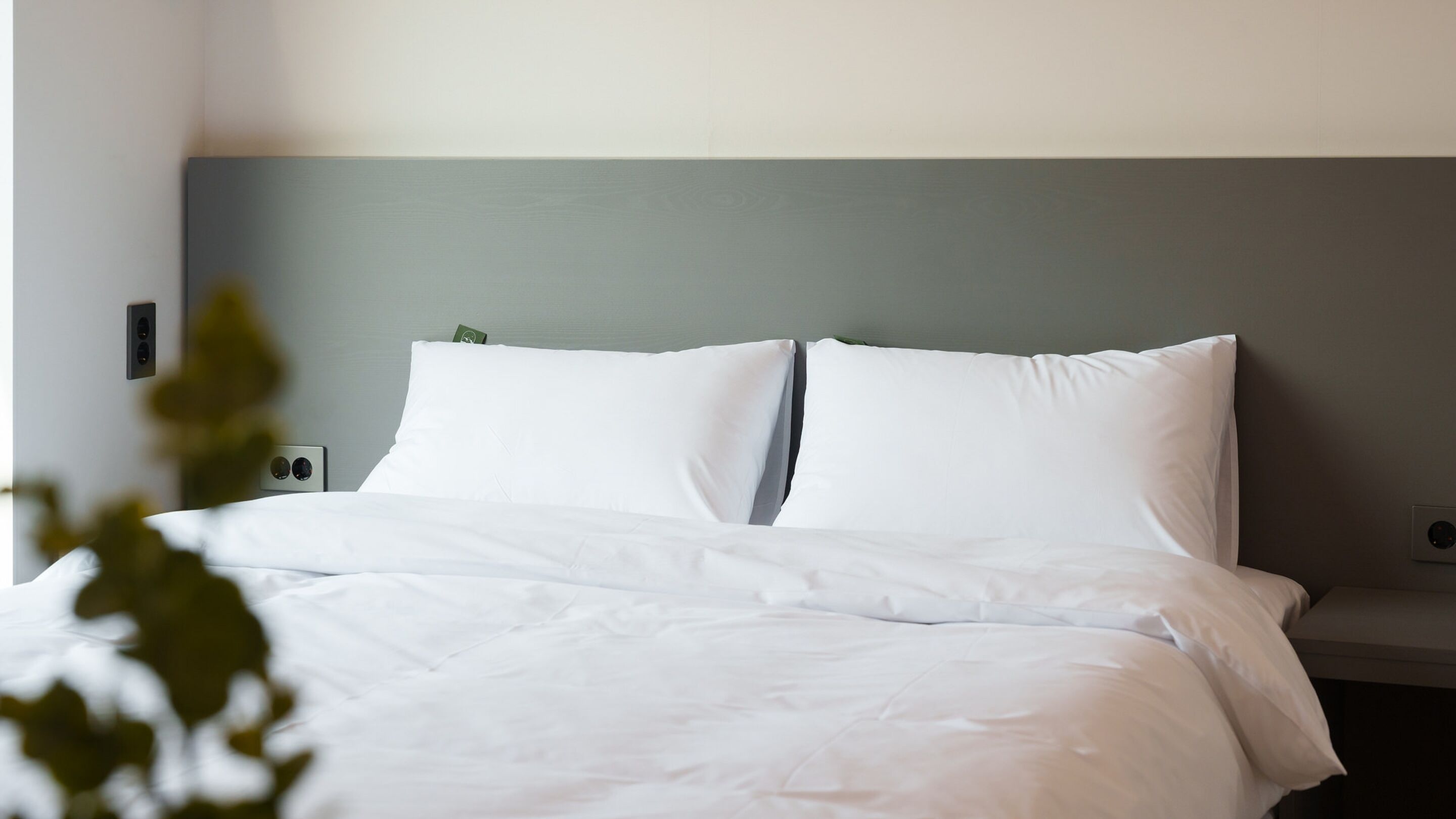 A neatly made bed with white linens, a grey headboard, and a small bedside table with a lamp.