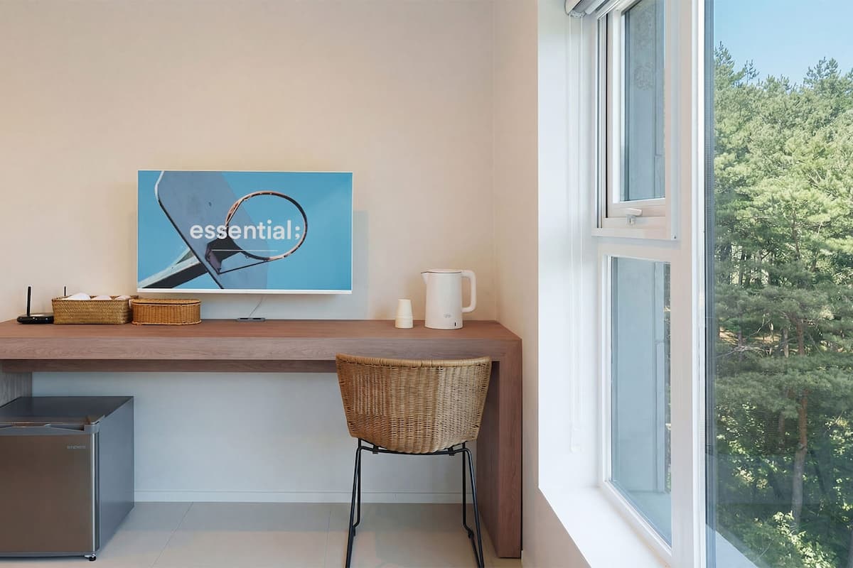 A modern hotel room with a wooden desk, a wicker chair, a small refrigerator, a coffee maker, and a framed artwork on the wall.