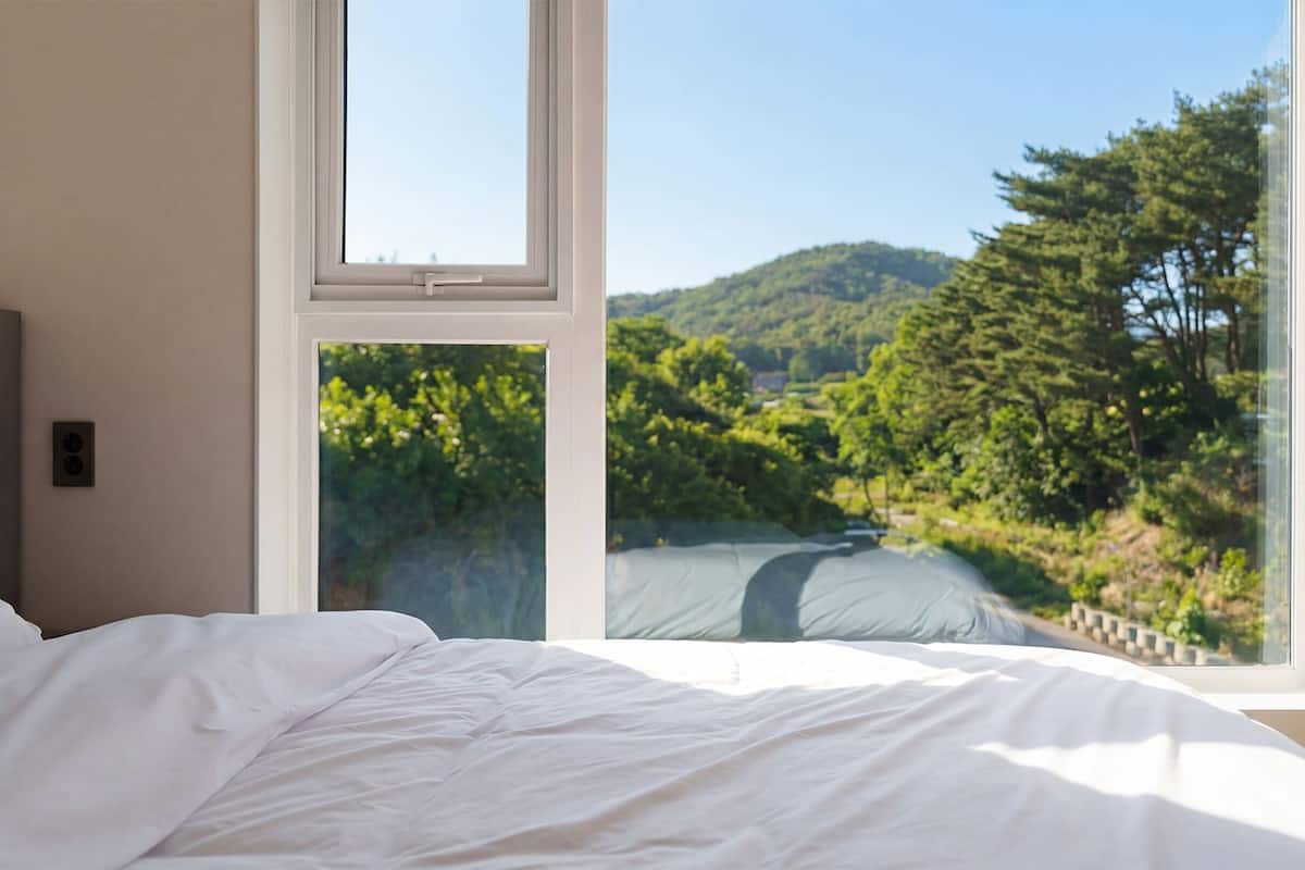 A bedroom with a bed and two windows offering a view of a forested hillside.