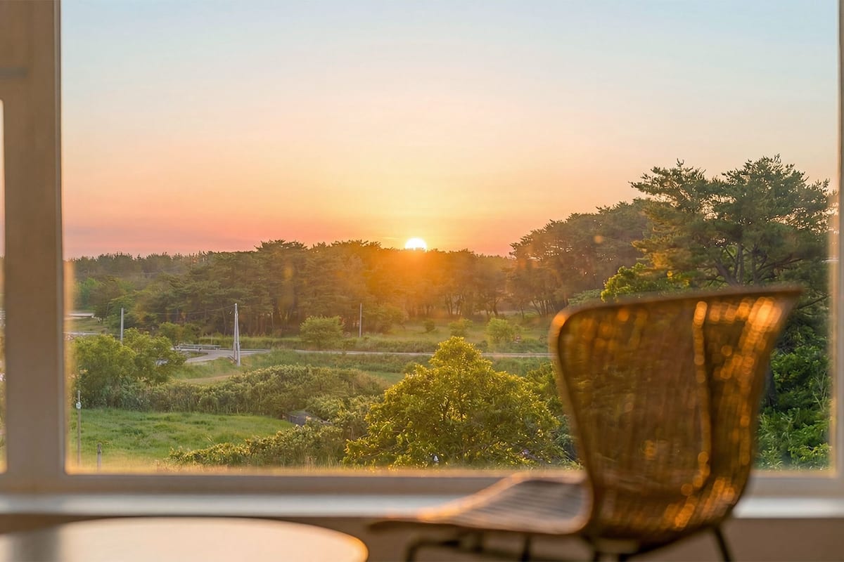 A room with a view of a sunset over a landscape, a chair, and a window.