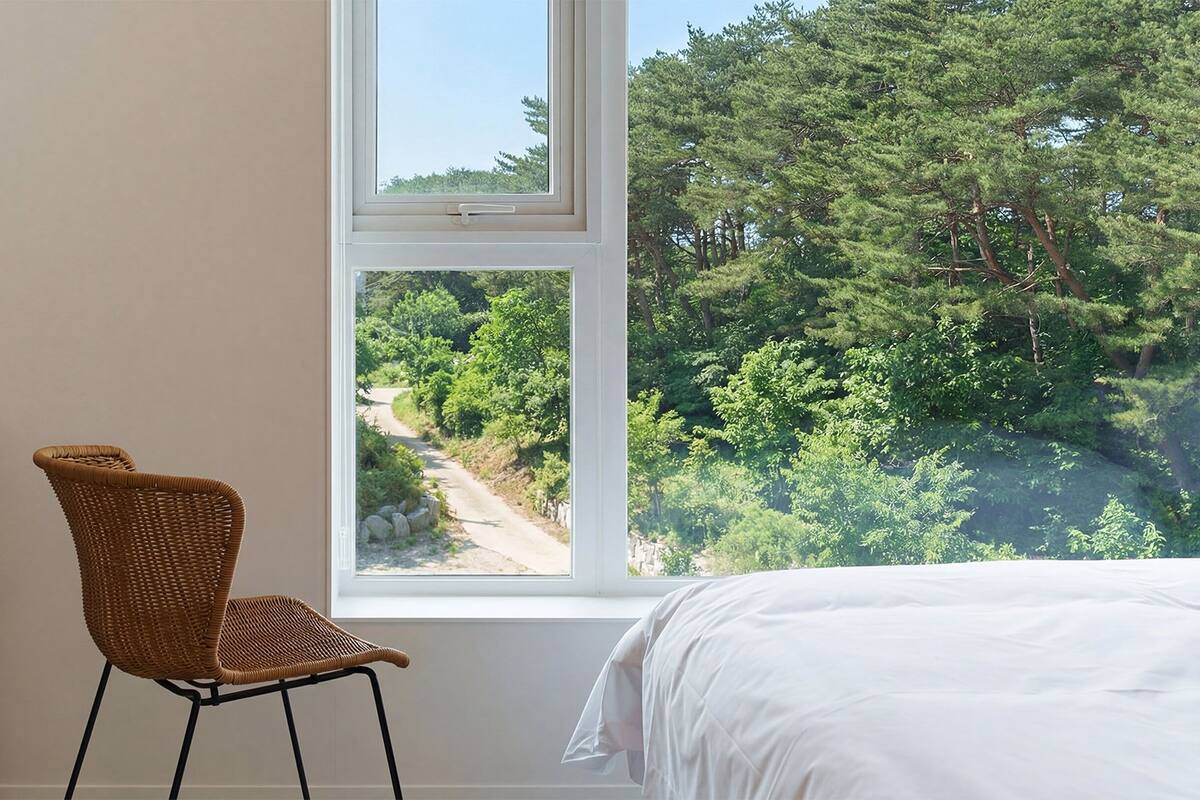 A bedroom with a large window, a wicker chair, a wooden desk, and a white bed.
