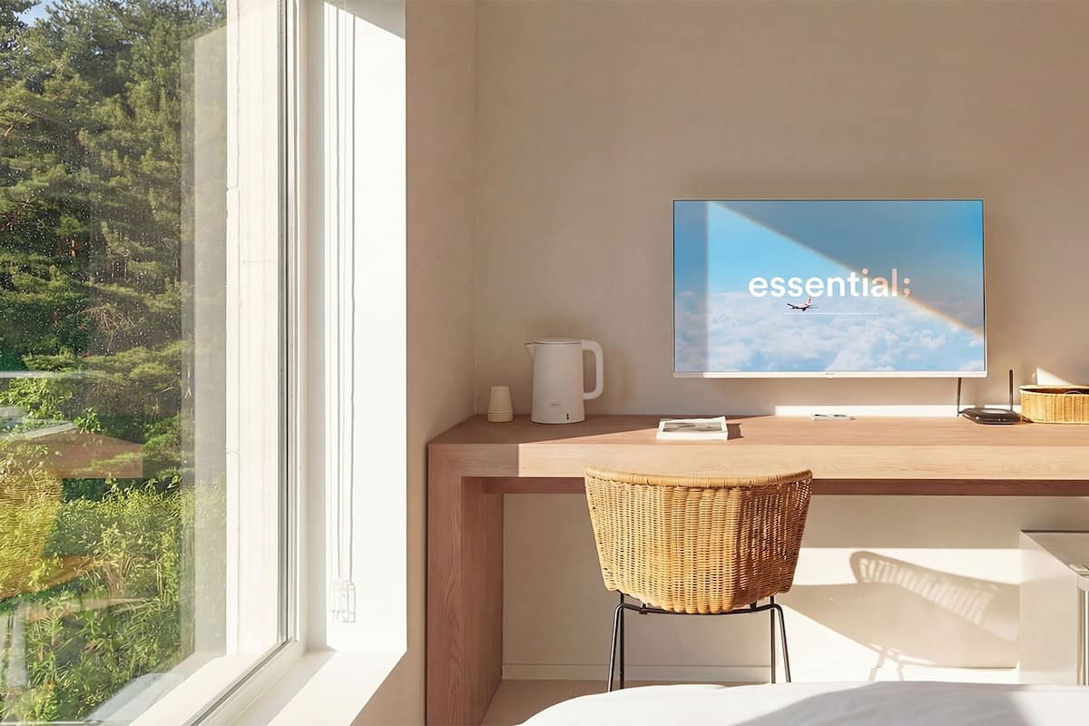 A modern hotel room with a large window, a flat-screen TV on a wooden desk, a wicker chair, and a bed.