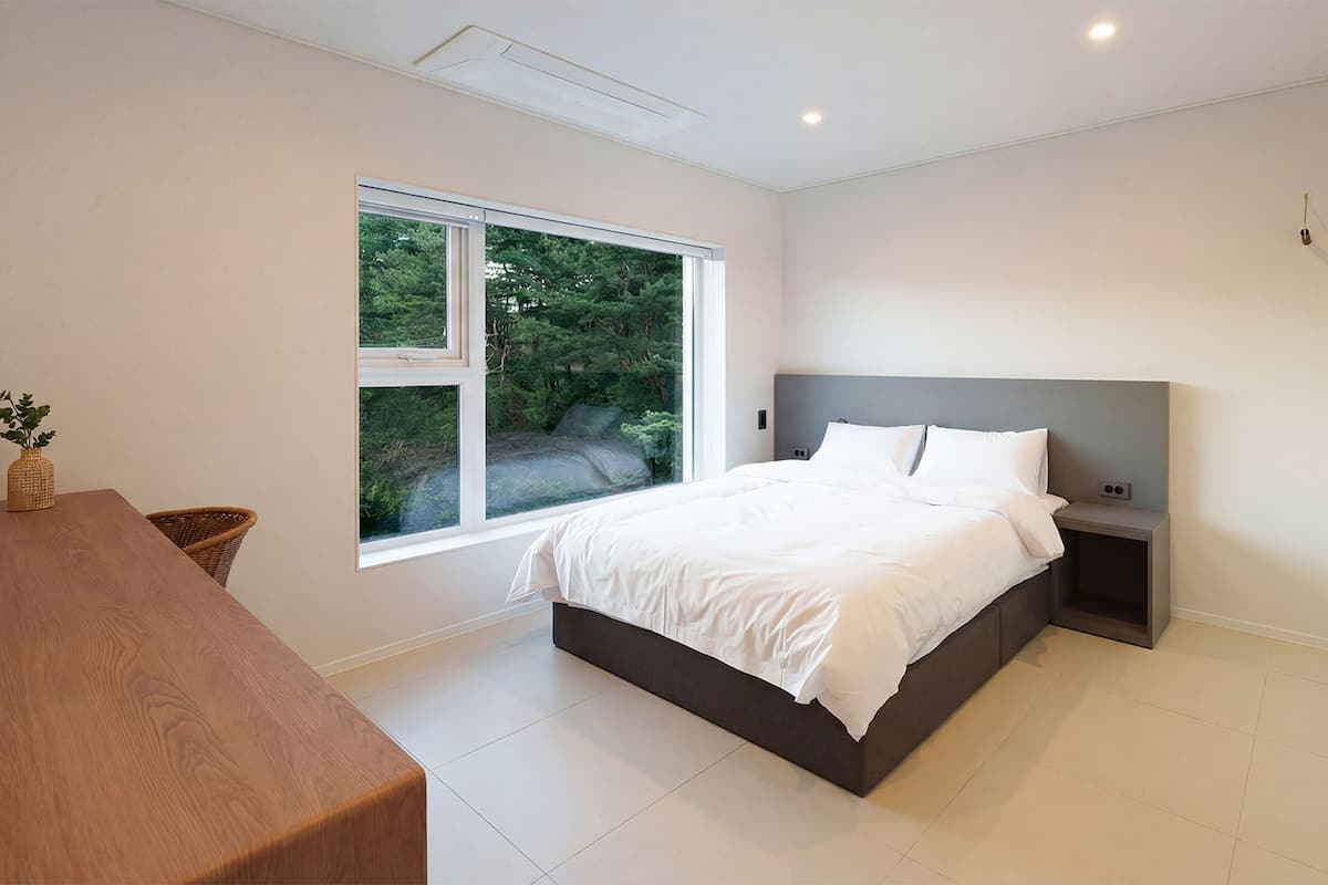 A modern bedroom with a large bed, a bedside table, a nightstand, a window with a view of greenery, and a wooden table.