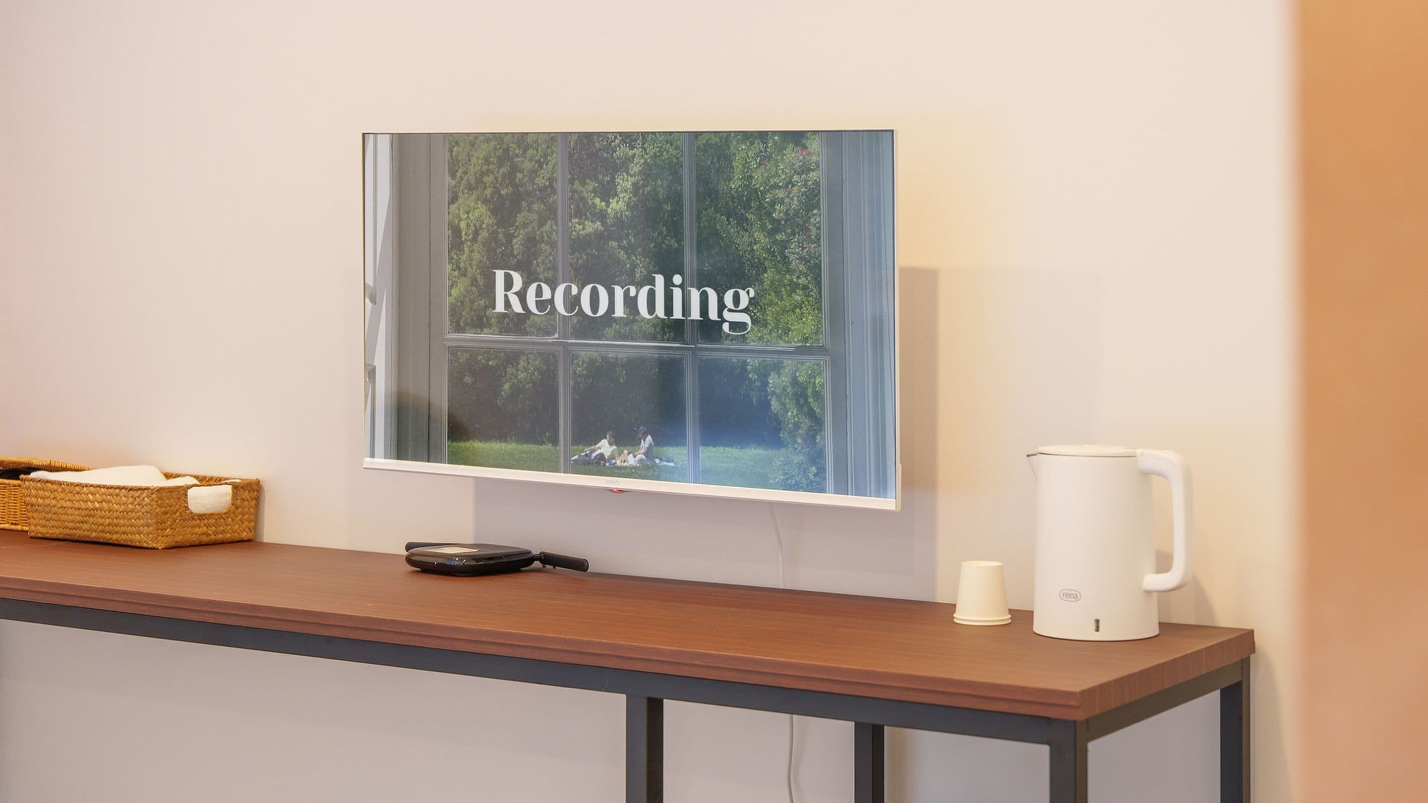 Ein Zimmer mit einem Holztisch, einer weißen Kaffeemaschine, einem Korb und einem Fenster mit der Aufschrift 'Recording'.