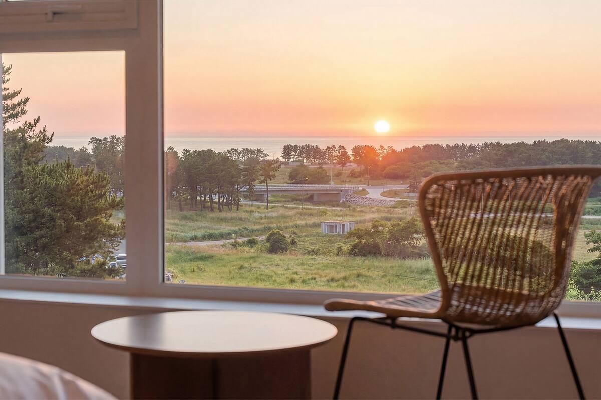 A room with a view of a sunset over a landscape, a wicker chair, and a small round table.