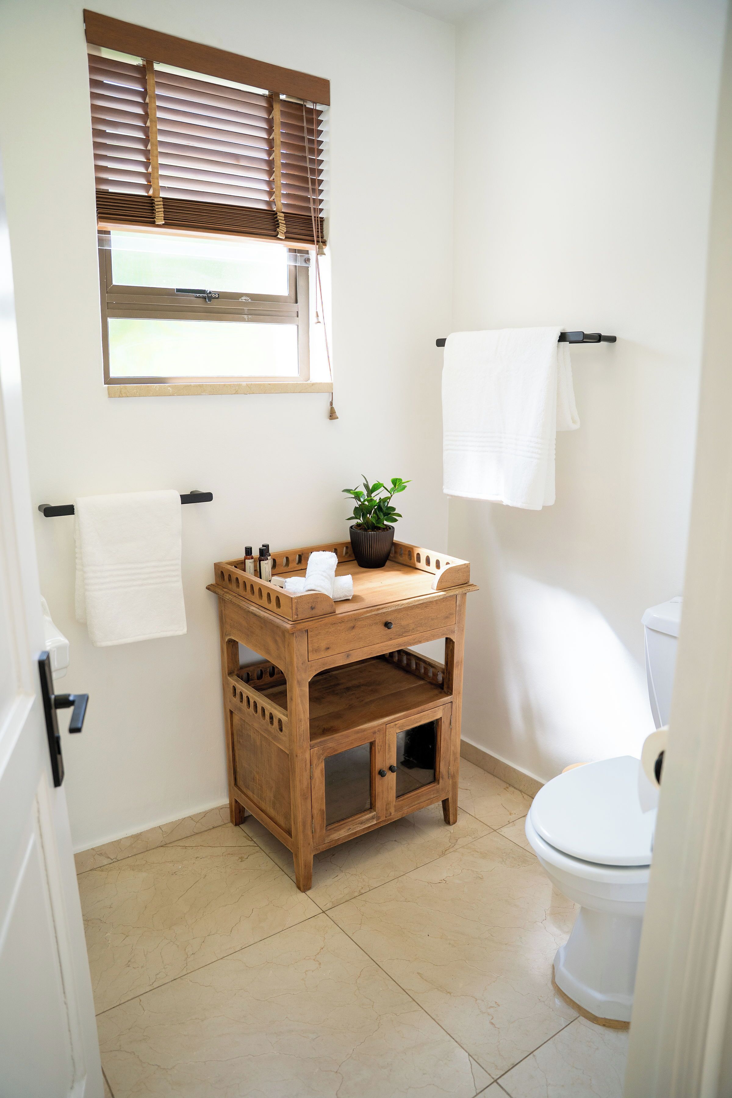 A bathroom with a wooden vanity, a toilet, a window with blinds, and a potted plant.