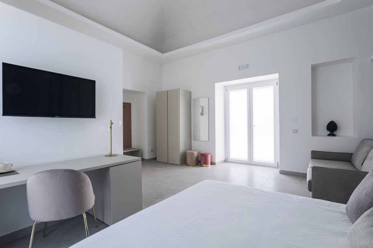Ein modernes Hotelzimmer mit einem großen Bett, einem Schreibtisch, einem Sessel, einem Fernseher, einem Kleiderschrank und einer Tür zu einem Balkon.