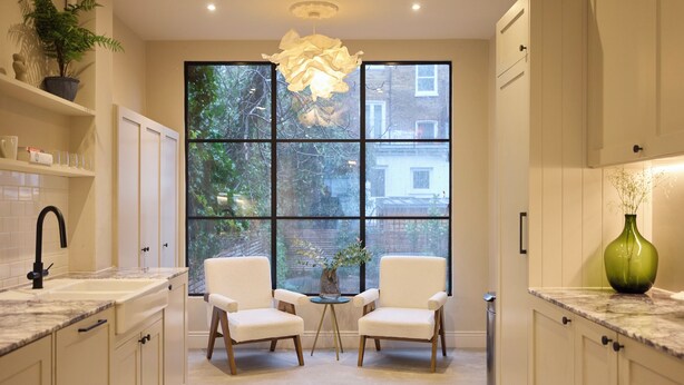 A modern kitchen with white cabinetry, a marble countertop, two white chairs, a small table with a vase, and a large window with a view of greenery and buildings. - The Architect's House (London)