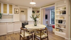 A modern kitchen with white cabinetry, a round dining table with wicker chairs, and a bookshelf with books and decorative items. - The Architect's House (London)