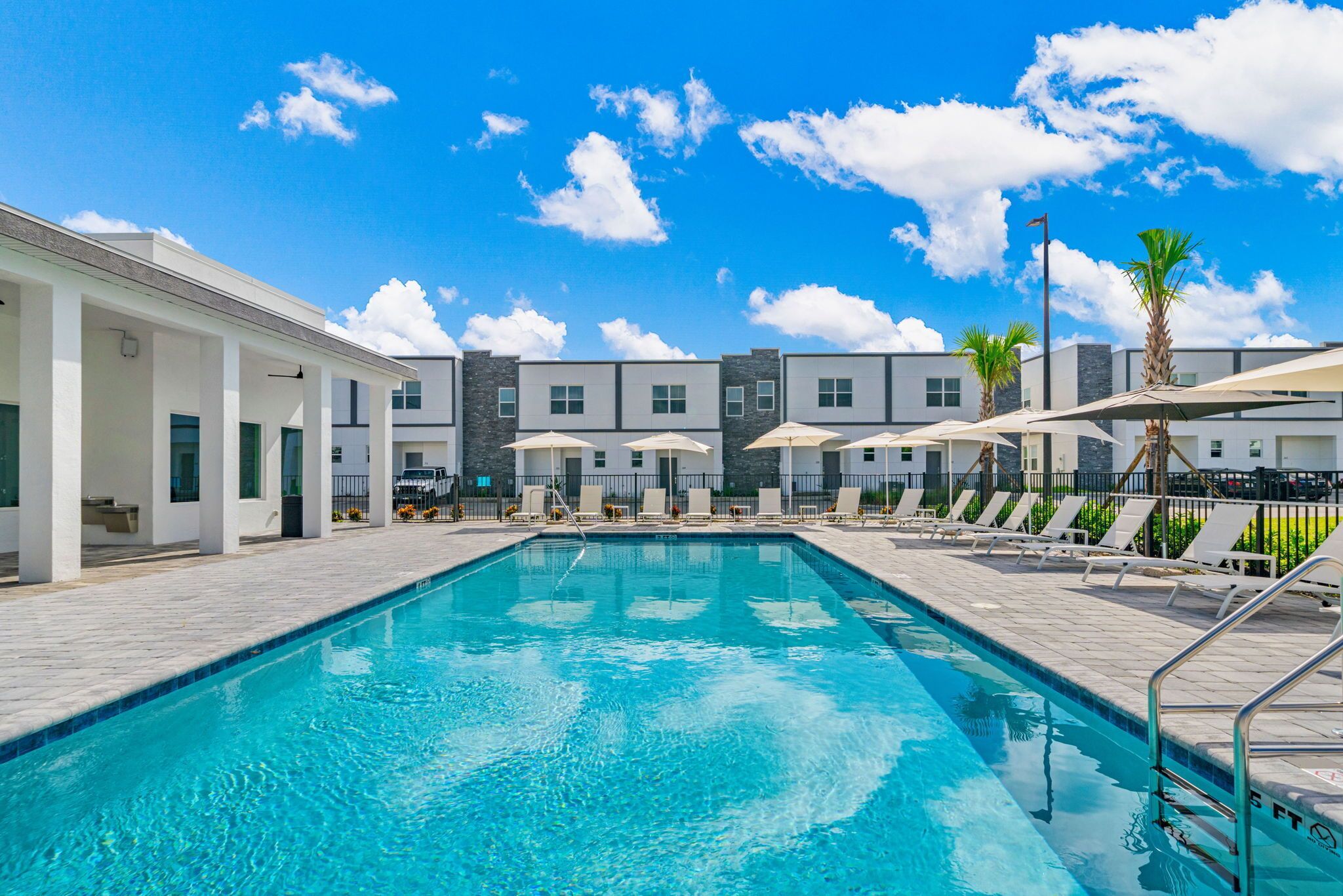 A swimming pool with lounge chairs and umbrellas, surrounded by modern buildings with a clear blue sky.