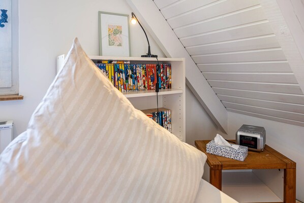 Un dormitorio con techo inclinado, una mesita de noche con un reloj y pañuelos de papel, una estantería llena de libros coloridos, y un almohadón grande.