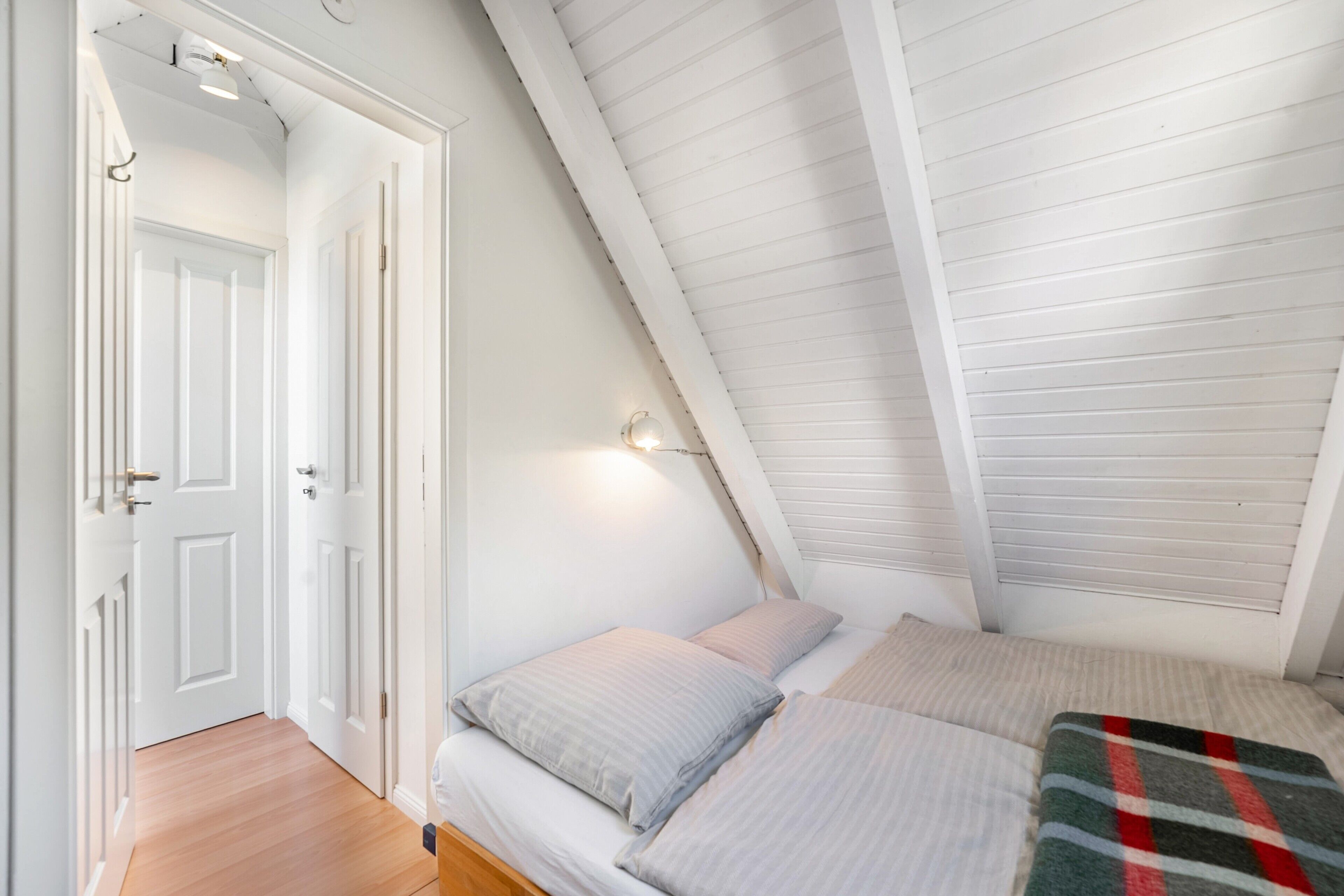 Une chambre avec un plafond en pente, un lit avec des oreillers et une couverture, et une porte donnant sur une autre pièce.