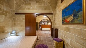 Ein Zimmer mit einer Steinswand, einem Bett, einem violetten Kissen, einem Gemälde und einem Bücherregal im Hintergrund.