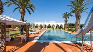 A pool area with a clear view of a building with arches in the background, surrounded by palm trees and a covered seating area with chairs. - Villa Arkè (Capilungo)