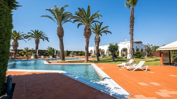 A pool area with palm trees, lounge chairs, and a building with arches in the background. - Villa Arkè (Capilungo)