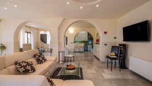 Ein modernes Wohnzimmer mit einer Couch, einem Couchtisch und einem Fernseher. Im Hintergrund ist eine Küche mit Essbereich zu sehen.
