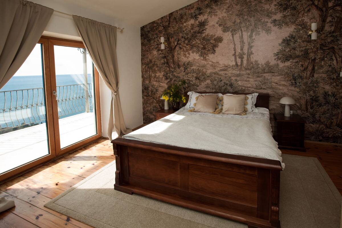 Une chambre avec un lit en bois, une vue sur la mer depuis un balcon et un papier peint sur le thème de la forêt.