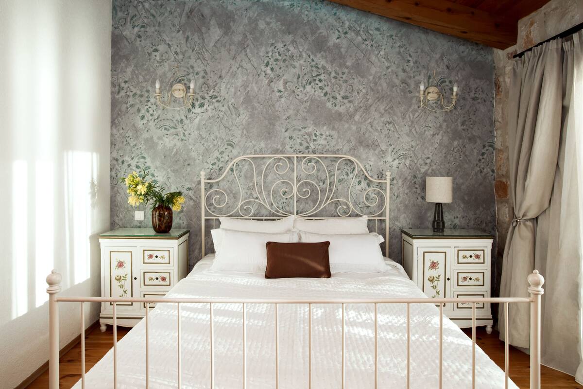 Une chambre à coucher avec un lit en métal, du linge de lit blanc, une table de chevet ornée d’un vase de fleurs et un mur à motifs texturés.
