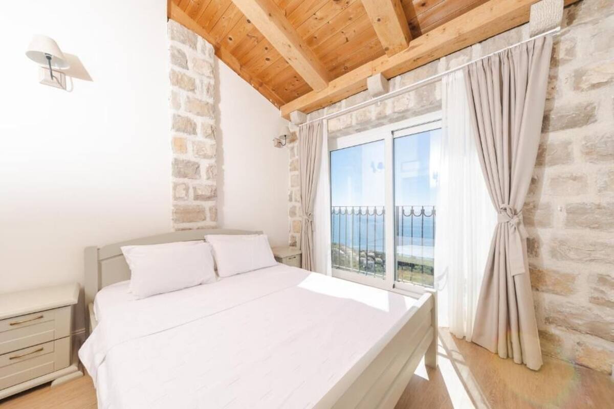 Une chambre à coucher comprenant un lit, une table de chevet, une lampe, un mur en pierre, un plafond en bois et un balcon avec une balustrade.